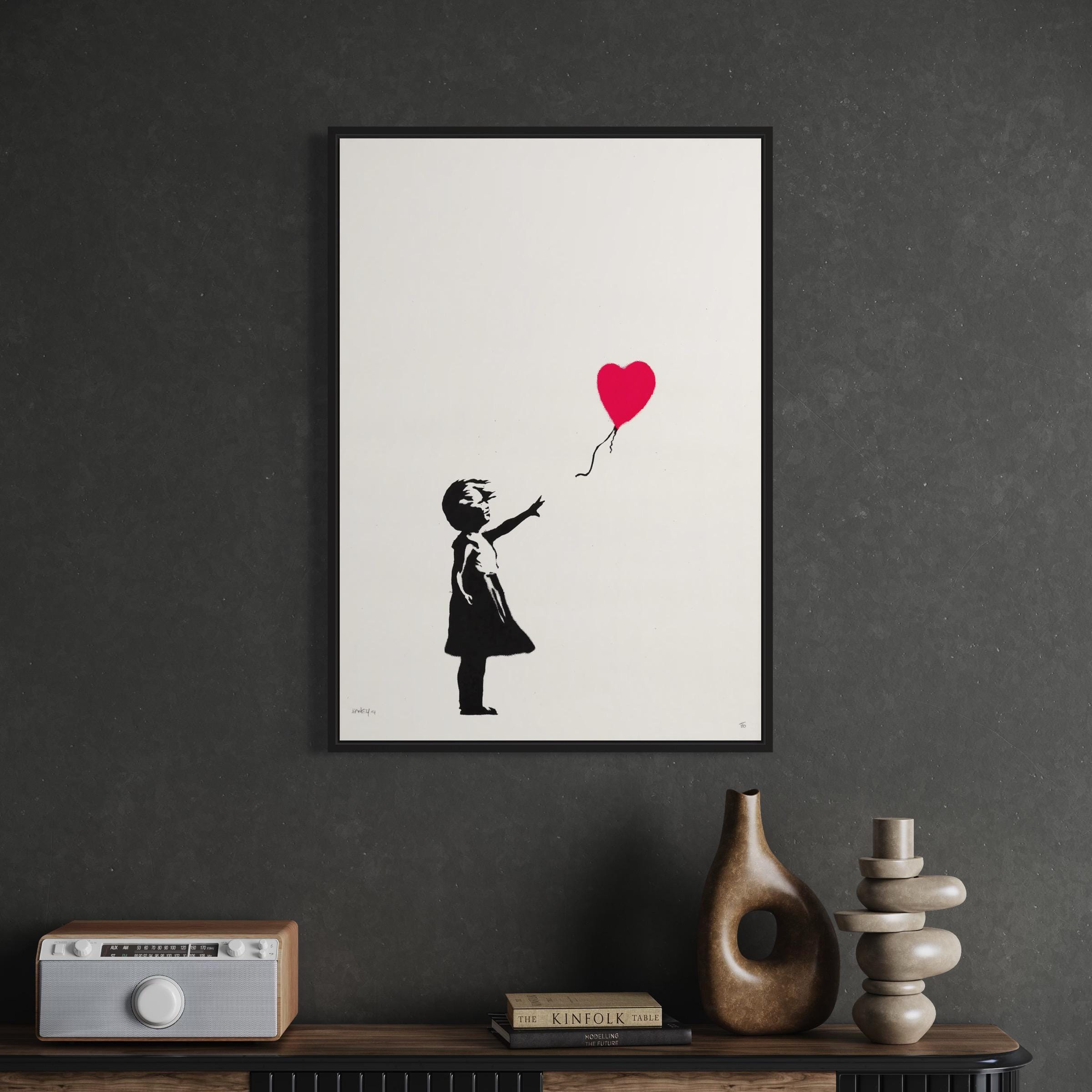 May include: A black and white stencil print of a girl reaching for a red heart shaped balloon. The print is framed in a black frame and is hanging on a dark gray wall.
